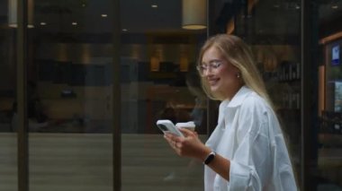 Genç, şık, gülümseyen, gözlüklü, caddede yürüyen, kahve içen, cep telefonu kullanan bir kadın. Millennial sarışın kadın, gözlüklü caddede yürüyor, kahvesini yudumluyor, telefonla mesajlaşıyor.