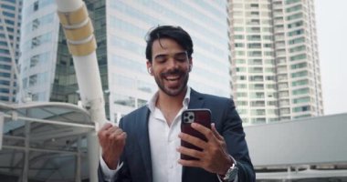 Ortadoğulu girişimci akıllı telefon ekranlı metropol caddesine bakarken neşe saçıyor. Geniş gülümseme ve jestler başarılı bir şekilde sonuçlanan anlaşmanın samimi neşesini ifade eder. Beklenmedik sevinç yüzünü aydınlatıyor