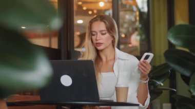 Sarışın, beyaz bir kadın güzel bir kafede dizüstü bilgisayarla çalışıyor yemyeşil bitkilerle telefonda konuşuyor. Sarışın genç bayan bilgisayar üzerinde çalışıyor modern cafe yemyeşil bitkiler cep telefonuyla konuşuyor