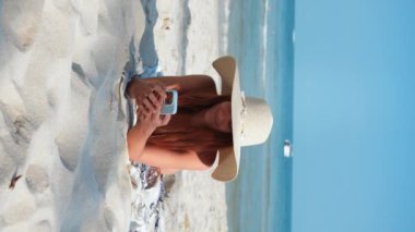 Dikey videolu kadın plaj tropiklerinde güneşleniyor ve cep telefonu kullanarak sohbet ediyor. Milenyum, sohbet uygulaması kullanılarak yazışırken deniz ve plajın güzelliğinin tadını çıkarıyor. Tatillerde iletişim kurmak için akıllı telefon kullanmak.