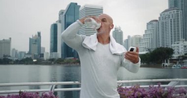 Beyaz bir adam antremandan sonra telefon ferahlatıcı sıvı kullanarak su şişesi içti. Enerjik şehir parkı. Olgun erkek sakin anı. İçme suyuna öncelik vermek için.