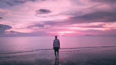 Kafkas erkek gezgin, akşam deniz üzerinde güzel bir mor gün batımının arkaplanına karşı sahilde yürüyor. Seyahat konsepti. Deniz tatili, egzotik sahil gezisi.