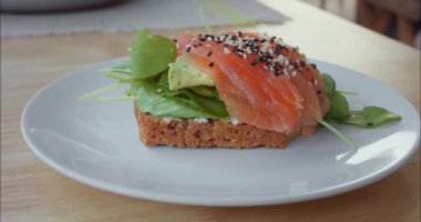 Kırmızı balık, susam, avokado, beyaz tabakta yeşil sandviç, ahşap masa. Mutfak yolculuğu, küresel tatların harmanlanması, epiküre yolculuğunu somutlaştırır. Yolculuğu kucaklamak lezzet yoluyla keşfetmek