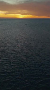 Deniz suyunda zarifçe yüzen teknenin dikey videosu görkemli turuncu günbatımı günbatımı deniz manzarası. Sakin okyanus sularında yüzen tekne canlı turuncu renkler gökyüzünü belirliyor