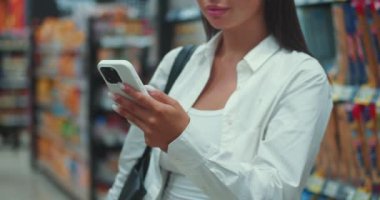 Genç yetişkin bir kadın cep telefonuyla, teknolojinin tüketici seçeneklerinde nasıl kilit rol oynadığını gösteriyor. Elinde telefon olan genç bir kadın modern teknolojiyi kullanarak ürünlerin arasında duruyor.