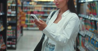 Geniş bir süpermarketin ortasında genç bir kadın teknoloji entegrasyonunu günlük tüketimde kullanıyor. Milenyum hanımı cep telefonunu kullanıyor modern teknoloji tüketiciler için bolluk çağını güçlendiriyor