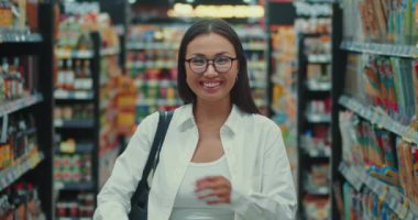 Kolları çaprazlanmış ve süpermarkette gülümseyen gözlüklü güzel bir genç kadın. Gözlüklü bin yıllık kadın süpermarkette kolları bağlı duruyor ve tüketici dünyasında gülümseme seçimi yapıyor.