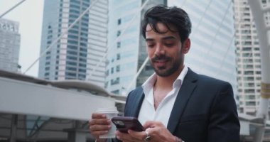 Takım elbiseli gülümseyen adam teneffüste sokakta kahve içer. Adam cep telefonunu elinde tutuyor ve sosyal medya yayınlarını inceliyor. Adam, etrafındaki gökdelenlere bakmak için alete ara veriyor.