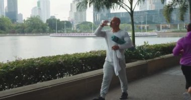 Atletik adam şehir parkının setinde yürüyor yoga minderiyle antrenman yaptıktan sonra dinleniyor ve su içiyor. Rahatlamış ve yürümekten ve gücünü toplamaktan zevk alan bir adam. Verimli derslerden sonra rahat yürüyüş.