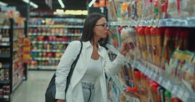 Süpermarkette duran gözlüklü güzel bir kadın birçok seçenek arasından en uygun olanı seçiyor. Gözlüklü çarpıcı kadın alışverişi düşünüyor süpermarkette geziniyor, seçenekleri değerlendiriyor.