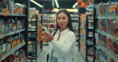 Çekici kadın, süpermarket tüketiciliğinde ve çeşitli ürünlerde kameraya gülümsüyor. Genç yetişkin bir bayan mağazada kamera ürünlerine gülümsüyor.