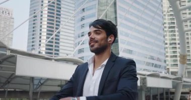 Resmi takım elbiseli ve kablosuz kulaklıklı tatmin olmuş yönetici, finans bölgesinde gülümsüyor. Kollar göğsün üzerinde ve gülümseme iyi ruh halini gösteriyor. Gökdelenlerin arasında dostça bir gülümseme davetkar görünüyor.