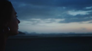 Alacakaranlıkta dinginliği kucaklayan bir kadın. Genç bir kadın, tropikal sahilde gün batımını canlı bir şekilde seyrediyor, doğanın çarpıcı güzelliğinden ilham alıyor ve huzur ve yansıma anından ilham alıyor.