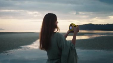 Huzurlu yaratıcı odaklanma anı. Genç kadın fotoğrafçı kumsalda gün batımının güzel resimlerini çekiyor kompakt dijital kamera kullanarak huzurlu atmosferin tadını çıkarıyor ve gökyüzünün çarpıcı renklerini yakalıyor.