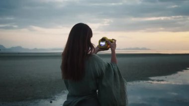 Altın saat boyunca plaj fotoğrafçılığı. Sahilde gün batımının güzel fotoğraflarını çeken kadın fotoğrafçı sarı kamerayla sakin bir anı fotoğrafçılık becerisiyle yakalıyor. Gün batımında plaj