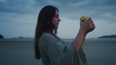 Plajda geçmişe dönük kamerası olan bir kadın. Genç bir kadın alacakaranlıkta sahilde duruyor, sarı aksiyon kamerasıyla fotoğraf ya da video çekiyor, sönen ışığın sakin atmosferinin tadını çıkarıyor.