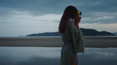 Gezgin blogcu manzarayı yakalıyor. Genç kadın fotoğrafçı, tropikal plajda güzel bir günbatımının fotoğraflarını çekiyor, klasik kamerayla, huzurlu atmosferin tadını çıkarıyor ve anı yakalıyor.