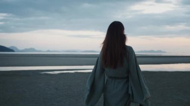 Gün batımında güzel kumsalda yürüyen genç bir kadın, huzurun, doğanın ve anın özgürlüğünün tadını çıkarıyor, arka planda dağlar ve sakin sular gökyüzünü yansıtıyor. Gün batımında plaj konsepti