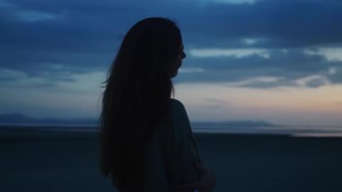 Alacakaranlık Gökyüzüne Karşı Siluetler. Uzun saçlı genç bir kadın plajda huzurlu bir gün batımının keyfini çıkarıyor. Yüzü izleyicilerden uzakta, engin bir okyanus ve sakin bir zemin oluşturan bir ufuk çiziyor.