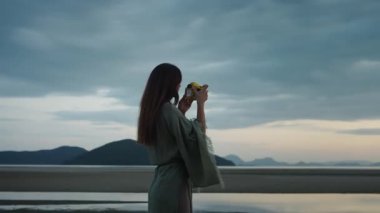 Dikkatli seyahat ve dijital anılar. Genç bayan fotoğrafçı plajda güzel gün batımı manzarası çekiyor, dingin atmosferi yakalıyor ve eski model kamerasıyla nefes kesici manzarayı yakalıyor.