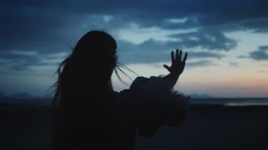 Gün batımında kumsalda özgürlüğünü ve mutluluğu kucaklayan genç bir kadının silueti. Rüzgar neşeyle saçlarını dalgalandırıyor ve kıyafetlerini dalgalandırıyor. Karamsar gökyüzünün altında kadınsı enerji