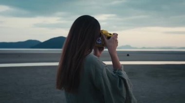 Sarı kamerayla fotoğraf çeken bir kadın. Genç kadın fotoğrafçı plajda büyüleyici günbatımı görüntüleri yakalıyor. Arka planda görkemli dağlar yükselirken, ellerinde parlak sarı bir kamera tutuyor.