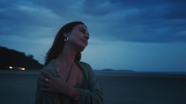 Boho şıklığında giyinmiş genç bir kadın kendini kucaklarken sakin, tenha bir plajda, doğa huzuruyla çevrili dramatik, bulutlu bir günbatımı gökyüzünün tadını çıkarıyor. Alacakaranlıktaki kadın