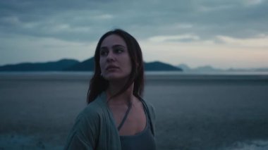 Özgürlük ve kendini ifade etme. Gün batımında sakin kumsalda yürüyen genç bir kadın, omuzlarının üzerinden bakarken, bulutlu gökyüzünün altında dağlar yükselirken, huzurlu bir yalnızlık anını yakalıyor.