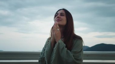 Kadın dışarıda minnettarlık pratiği yapıyor. Dua eden ellerini sıkan genç bir kadın, yüzü göğe yükselmiş, huzur dolu, açık havada, bulanık dağlar ve deniz manzaralı bir sükunet içinde.