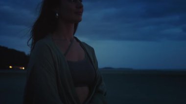 Gün batımı siluetleri ve sükunet. Şala sarılmış ve spor sutyen giyen bir kadın, sakin gölün üzerinde dramatik alacakaranlığın tadını çıkarıyor, gün batımına doğru yavaşça kaybolan huzurlu atmosferi kucaklıyor.