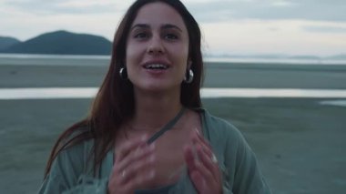 Yavaş Yaşam ve Dijital Detox. Uzun kahverengi saçlı ve yeşil ceketli genç bir kadın, arka planda dağlarla güzel bir sahilde günbatımının tadını çıkarırken gülümsüyor ve uzaklara bakıyor.