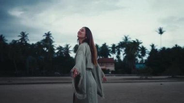 Gün batımında meditasyon dansında, uzun kahverengi saçlı, zarif yeşil elbiseli bir kadın kollarını kaldırıyor ve arka planda palmiye ağaçları ve okyanusla kaplı tropikal sahilde bulutlu gökyüzünün keyfini çıkarıyor.