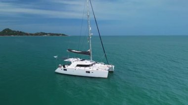 Siyah yelkenli modern beyaz katamaran, turkuaz deniz kıyısında, yeşil kıyı şeridinde, güzel bulutlu gökyüzünün altında yaz tatilinin tadını çıkarıyor. Tropikal dinginlikte yüzen katamaran