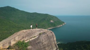 İki turist, Tayland 'da doğanın sükunetini kucaklarken sakin bir körfez ve engin turkuaz okyanusa bakan kayalık zirveden nefes kesici manzaranın keyfini çıkarıyorlar. Çift kayalık zirvenin kenarında duruyor.