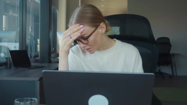 Finansal analist kadın, modern ofiste dizüstü bilgisayarla çalışırken baş ağrısı çekiyor, elleriyle alnına dokunuyor, yorgunluk ve hüsranı gösteriyor. Ofiste yorgunluk.