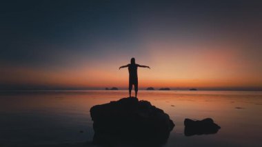 Parlayan ufka bakan açık kollar. Kolları açık kayaların üzerinde duran turist silueti, sakin tropikal ada sahillerinde sakin okyanusun üzerinde canlı günbatımının tadını çıkarıyor, dingin ve ilham verici bir seyahat sahnesi yaratıyor.