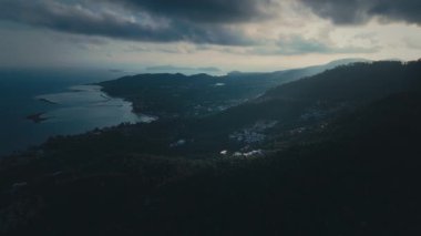 Bereketli bitki örtüsünün kayan tepeleri kapladığı, kıyı boyunca yayılmış evler ve gün batımında gölgelerini düşüren dramatik bulutlu gökyüzü ile tropik kıyı şeridinin çarpıcı hava manzarası. Tropiklerde muson yağmurları