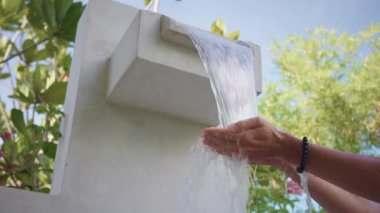 Turist ellerini modern açık hava lavabosunda yıkıyor, yemyeşil ve mavi gökyüzünün zeminine karşı ferahlatıcı temiz su akıntısının tadını çıkarıyor. Serinletici şelaleli açık hava duşu