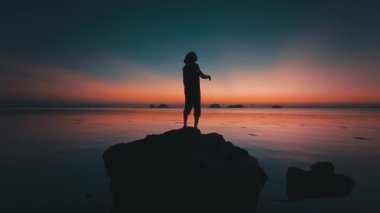 Kayalık kıyı şeridinden gün batımını izliyorum. El Nido, Palawan, Filipinler 'de sakin bir okyanusta kayaların üzerinde canlı günbatımının keyfini çıkaran küçük adalar görülebilen turist silueti.