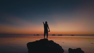 Adam gün batımında insansız hava aracı fırlatıyor. Tropikal günbatımını kutlayan turistlerin silueti deniz kenarında kaya oluşumunda dururken bir kolunu kaldırıp uzaktan kumandalı insansız hava aracını tutuyor.