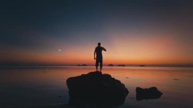 Parlayan dron insanın etrafında uçuşuyor. Kaya oluşumunun üzerinde duran turist silueti, sakin tropikal okyanusun üzerinde güzel bir günbatımını hayranlıkla seyrederek, uzak adalarda huzurlu ve huzurlu bir atmosfer yaratıyor.