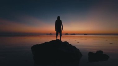 Gün batımında okyanusta kayaların üzerinde duran genç adamın silueti, gökyüzünün canlı renklerinin tadını çıkarıyor ve tropik cennetin huzurlu atmosferini kucaklıyor. Canlı günbatımında, kayanın üzerinde siluet.