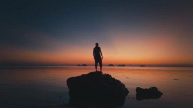 Adam altın alacakaranlıkta hareketsiz duruyor. Deniz kenarında kayaların üzerinde duran, gün batımının canlı renklerine bürünmüş, uzak adalarda akşam gökyüzüne karşı çarpıcı bir zemin oluşturan turist silueti.