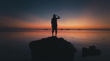 Yalnızlığın ardında parlayan gökyüzü. Okyanustaki kayaların üzerinde duran turist silueti sakin denizin üzerinde renkli günbatımının tadını çıkararak sakin ve huzurlu bir atmosfer yaratıyor. Okyanus yamacında gün batımı yalnızlığı