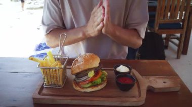 Turist ellerini ovalarken lezzetli hamburgerin tadını çıkarıyor. Taze malzemeler, çıtır patatesler ve Amerikan mutfağı olan sahil restoranındaki ahşap ahşap tahta üzerinde soslar.