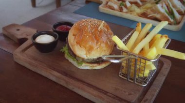 Susamlı hamburgerin yanında patates kızartması, ketçap ve mayonez servis edilir. Tahta bir tahtaya davetkar bir şekilde oturur ve tatmin edici bir yemek sözü verir. Yetersiz beslenme