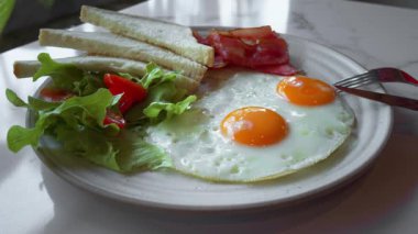 İki kızarmış yumurta, kızarmış pastırma şeritleri, kızarmış ekmek dilimleri ve kiraz domatesli taze salata beyaz tabakta dengeli ve cezbedici bir kahvaltı yaratır. Tam Amerikan Kahvaltısı