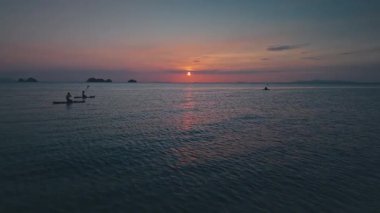 Güneş uzak adaların ardından batarken sakin okyanus sularında sakin kürek çekme seansının keyfini çıkaran üç turist, gökyüzü ve deniz boyunca sıcak bir ışık saçıyor. Alacakaranlık renkli denizi geçen yalnız bir tekne.