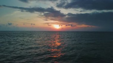 Güneş dingin okyanusun üzerinde batarken, su yüzeyinde sıcak altın parıltı oluştururken dramatik bulutlar gökyüzünü doldururken, pitoresk akşam atmosferini güçlendiriyor. Sakin deniz ufkunun üzerinde güneş görünüyor.