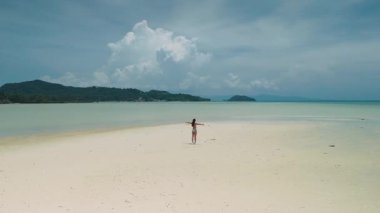 Geniş kolları olan el değmemiş beyaz kumsalda duran genç bir kadın yemyeşil adaları ve arka planda dramatik bulutları olan tropik bir cennetin sükunetini kucaklıyor.